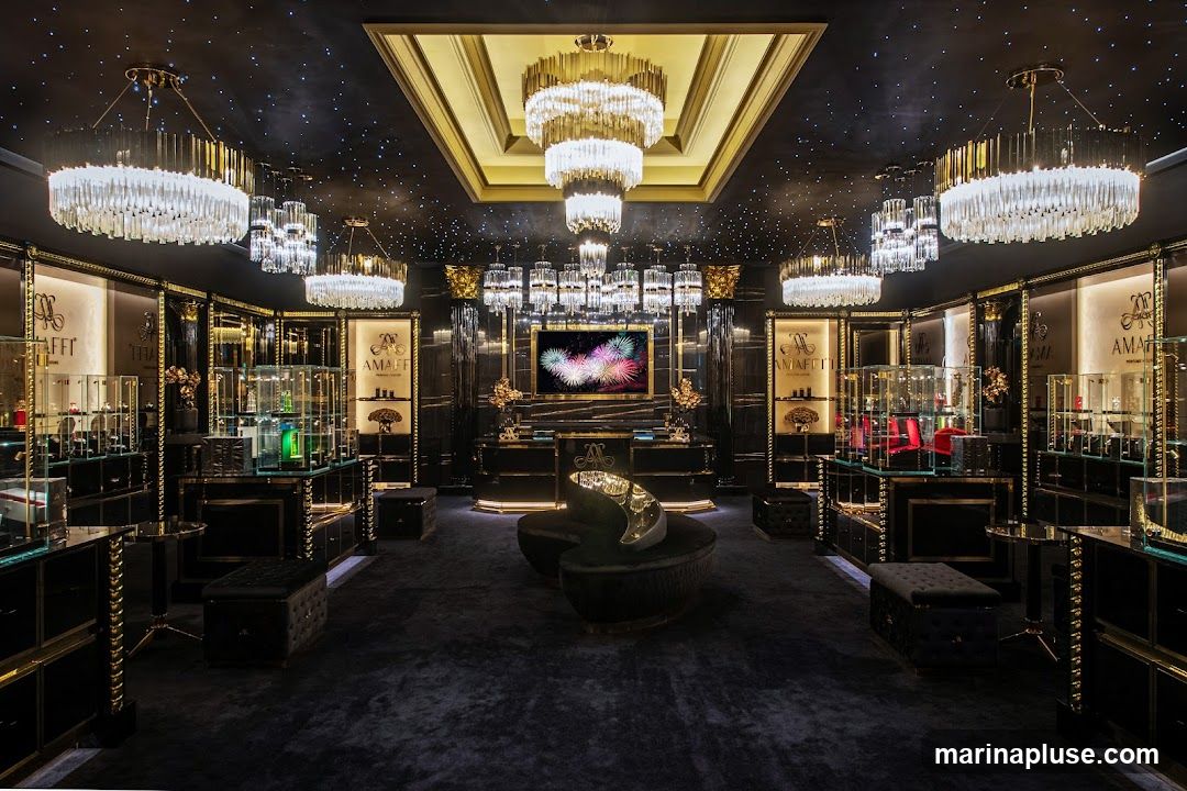 The interior of a high-end perfume store is depicted, featuring a dark, opulent design with gold accents. The ceiling is black with a starry effect and adorned with multiple elaborate crystal chandeliers. The walls are lined with glass display cases showcasing various perfume bottles and products, illuminated from within. In the center of the room, a modern, curved black velvet sofa with a metallic sculpture sits on a plush dark carpet. A large screen displays a vibrant fireworks display. The store's branding, "AMALFI PERFUME LONDON," is visible on illuminated panels behind the display cases.