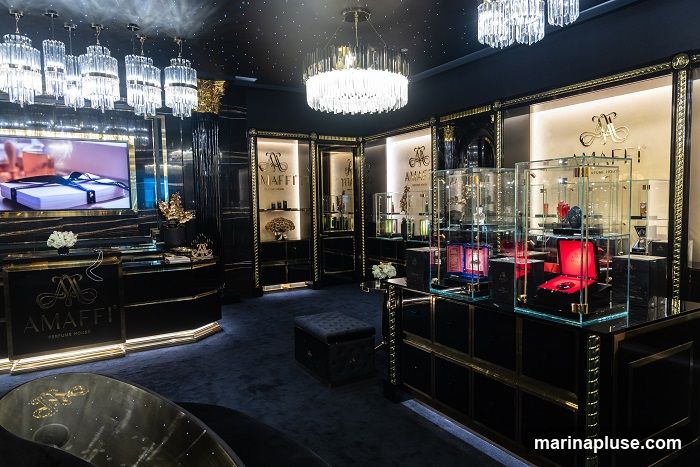 The interior of a high-end perfume store, featuring opulent decor. The walls and display cases are black with gold accents. Crystal chandeliers hang from the ceiling, and the floor is covered in dark blue carpet. Various perfumes are displayed in glass cabinets. A large screen shows an advertisement for a gift-wrapped product. The store's logo, "AMaffi PERFUME HOUSE," is visible on signage and displays.