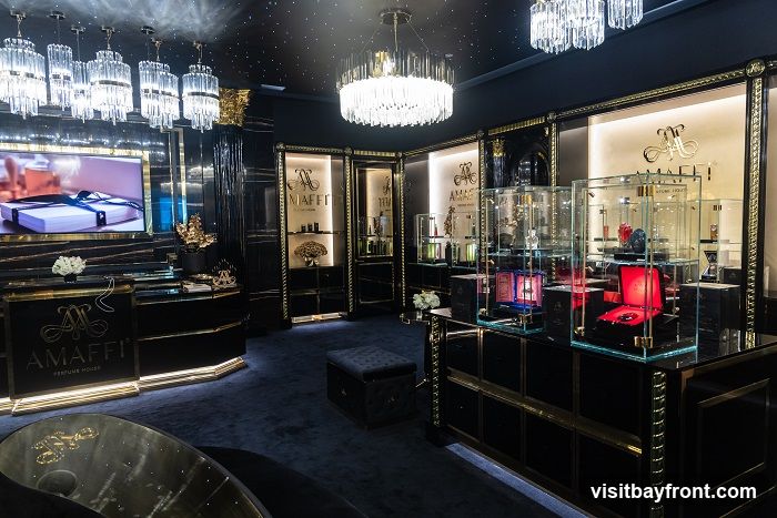The interior of a high-end perfume store, featuring opulent decor. The walls and display cases are black with gold accents. Crystal chandeliers hang from the ceiling, and the floor is covered in dark blue carpet. Various perfumes are displayed in glass cabinets. A large screen shows an advertisement for a gift-wrapped product. The store's logo, "AMaffi PERFUME HOUSE," is visible on signage and displays.