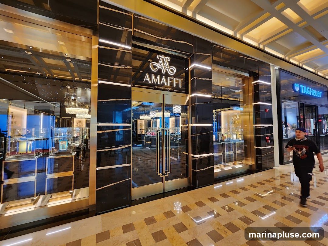 The exterior of a luxury perfume store with a black and gold facade. The store's name, "AMaffi Perfume House," is prominently displayed above the entrance. Inside, chandeliers and display cases filled with perfume bottles are visible. To the right, a TAG Heuer store is visible. A man walks past the storefront.