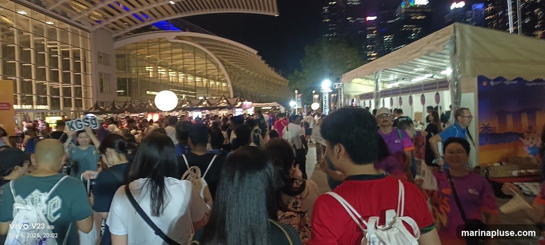 A large crowd of people are gathered outdoors at night, with a modern building structure in the background. There are tents and stalls set up, suggesting a market or festival. Some people are holding signs, and the atmosphere appears lively.