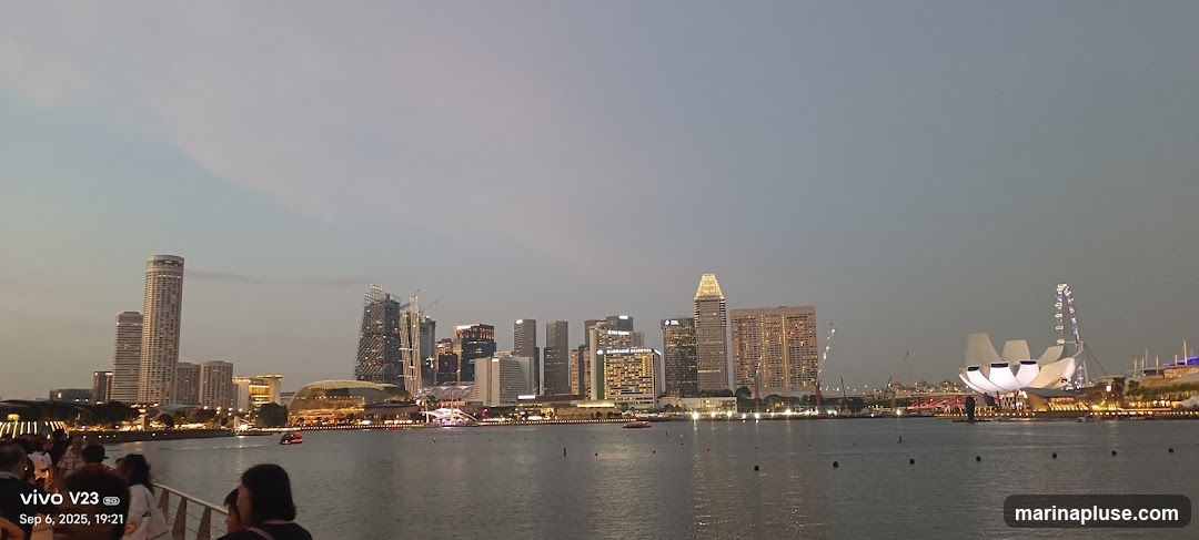 A panoramic view of the Singapore skyline at dusk, with various skyscrapers and the Marina Bay Sands complex visible across the water. Several boats are on the water, and people are gathered on a walkway in the foreground. The sky is a soft gradient of blue and grey.