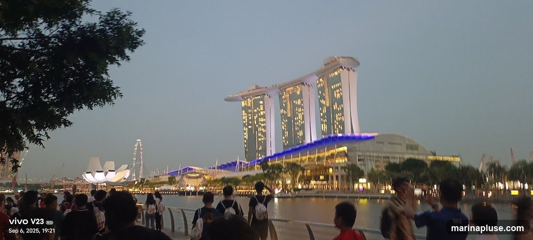A wide shot of the Marina Bay Sands complex in Singapore as dusk settles. The iconic three towers, connected by the SkyPark, are illuminated with warm lights. Below, the building's facade glows with a blue light strip. In the foreground, a crowd of people are gathered along the waterfront, some taking photos. A Ferris wheel is visible in the distance to the left, and a lotus-shaped building is also present. The sky is a soft, dusky blue.