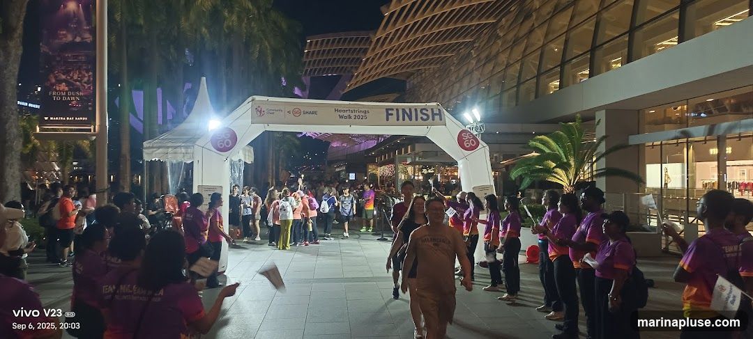 A crowd of people are gathered at a finish line archway for the Heartstrings Walk 2025. Some participants are crossing the finish line, while others are lined up on the sides, some holding small flags. The event appears to be taking place outdoors at night, with a modern building facade in the background. A banner above the archway reads "Heartstrings Walk 2025 FINISH". There are also signs indicating "SG 60" and logos for "Community Chest" and "SHARE". A large vertical banner on the left side of the image advertises an event called "FROM DUSK TO DAWN" at "MARINA BAY SANDS".
