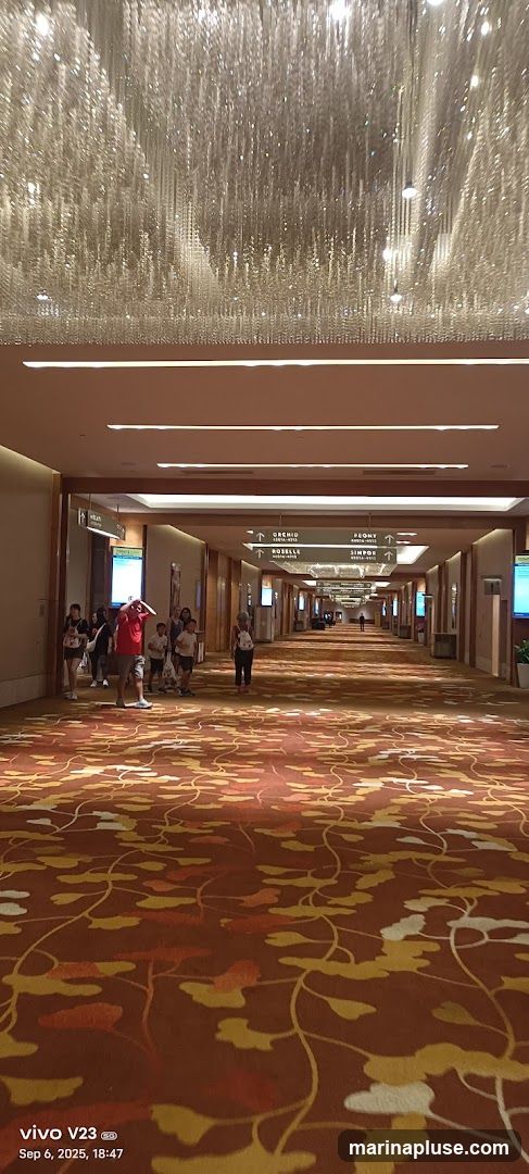 A long, wide hotel corridor with a highly decorative ceiling featuring a large, sparkling chandelier. The walls are lined with wooden panels and illuminated by recessed lighting. Several directional signs are visible overhead, indicating various room names such as 'ORCHID', 'PEONY', 'ROSELLE', and 'SIMPOR'. People are seen walking down the corridor. The floor is covered with a richly patterned carpet in shades of orange, red, and yellow, with a floral or leaf motif.