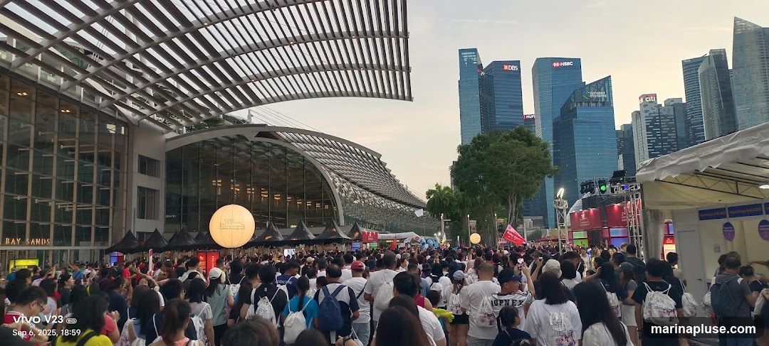 A large crowd of people are gathered outdoors in front of a modern building with a distinctive curved roof structure. In the background, several tall skyscrapers, including buildings with HSBC and DBS logos, dominate the city skyline. There are also tents and stage setups visible, suggesting an event or festival is taking place. A large illuminated sphere with text is also present. The sky is overcast with the warm tones of dusk.