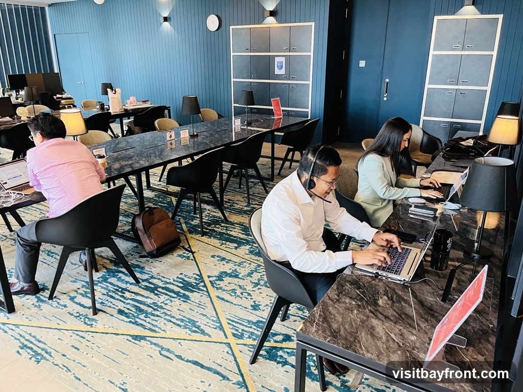 Several individuals are seated at a long, marble-topped desk in a contemporary office environment. The walls are painted a deep blue, and the floor is covered with a patterned carpet. Each person is engaged with a laptop, with some wearing headsets. The space is furnished with black chairs, desk lamps, and shelving units.