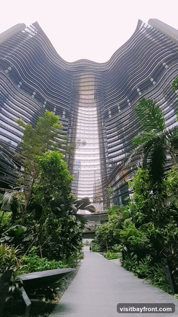 A pathway leads through a lush, tropical garden situated within a modern architectural marvel. The buildings surrounding the garden feature a distinctive wavy, layered facade with numerous windows. The sky is overcast, creating a soft, diffused light. A lone figure walks in the distance on the pathway.