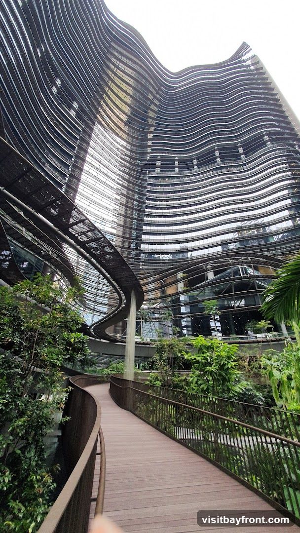 A walkway curves through a verdant indoor garden, with lush green plants and trees on either side. A waterfall cascades down from an elevated structure. In the background, a striking modern building with a wavy, layered facade rises high into the sky. The building's exterior is composed of numerous horizontal bands of glass and dark material, creating a dynamic, undulating appearance.