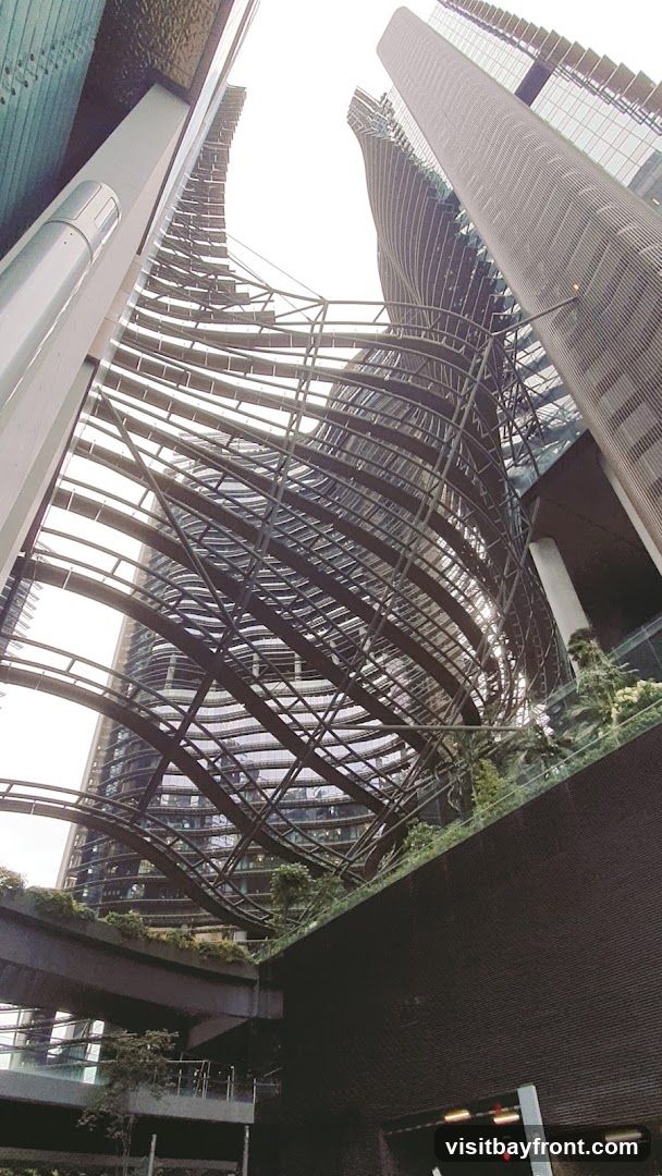 A low-angle shot looking up at the complex, modern architecture of several skyscrapers. The buildings feature curved facades, intricate metal frameworks, and extensive glass windows. Greenery is visible on elevated platforms and terraces integrated into the structures. The sky is overcast.
