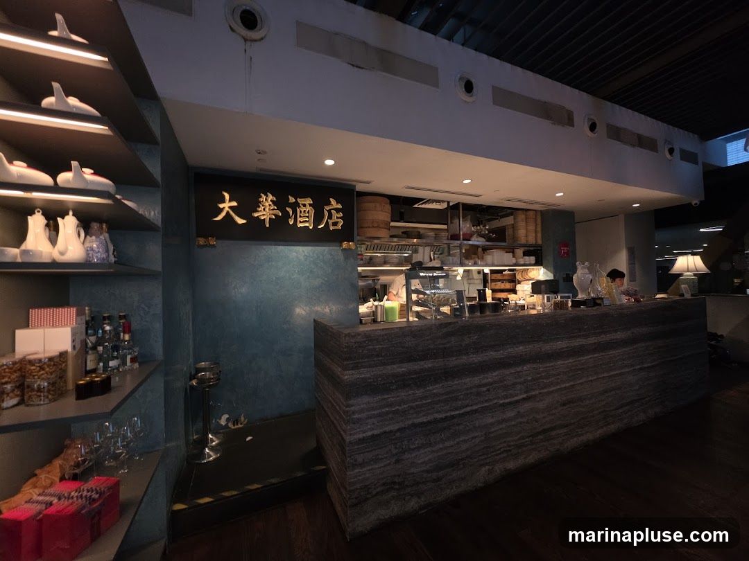 A view of a restaurant counter with a marble-like facade. Behind the counter, kitchen staff are visible preparing food. Shelves to the left display decorative items and bottles. A sign with Chinese characters is mounted on the wall above the counter.