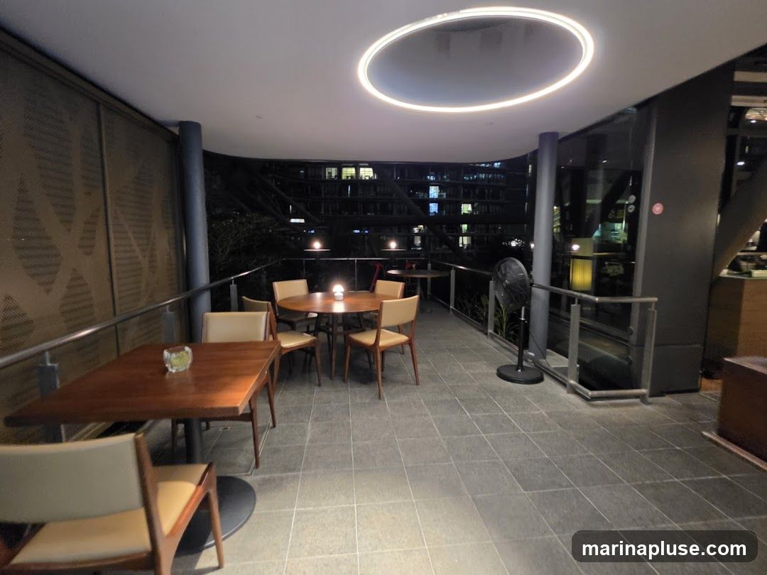 An outdoor seating area with tables and chairs is shown at night. The area is paved with large gray tiles and features a railing along the edge. In the background, buildings with illuminated windows are visible. A circular light fixture is mounted on the ceiling. A standing fan is placed near the railing.