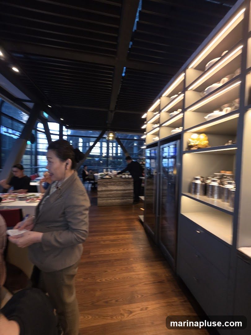 A woman in a grey blazer stands in a restaurant with wooden floors. To her right is a tall shelving unit with illuminated shelves displaying white teapots and other decorative items. In the background, other diners are seated at tables, and a server is visible near a counter. The restaurant has large windows and a modern industrial-style ceiling.