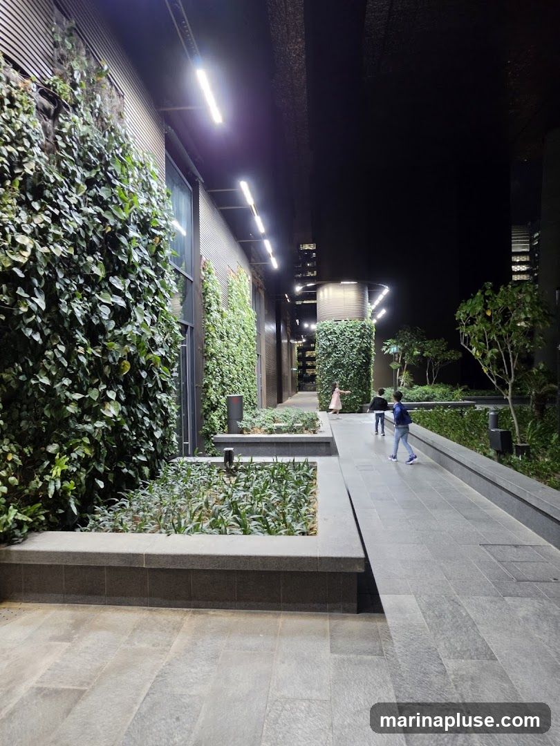 A nighttime view of a modern outdoor walkway featuring lush vertical gardens on the side of a building. The walkway is paved with large, grey stone tiles. Several people are walking along the path. Linear lights illuminate the scene from above.