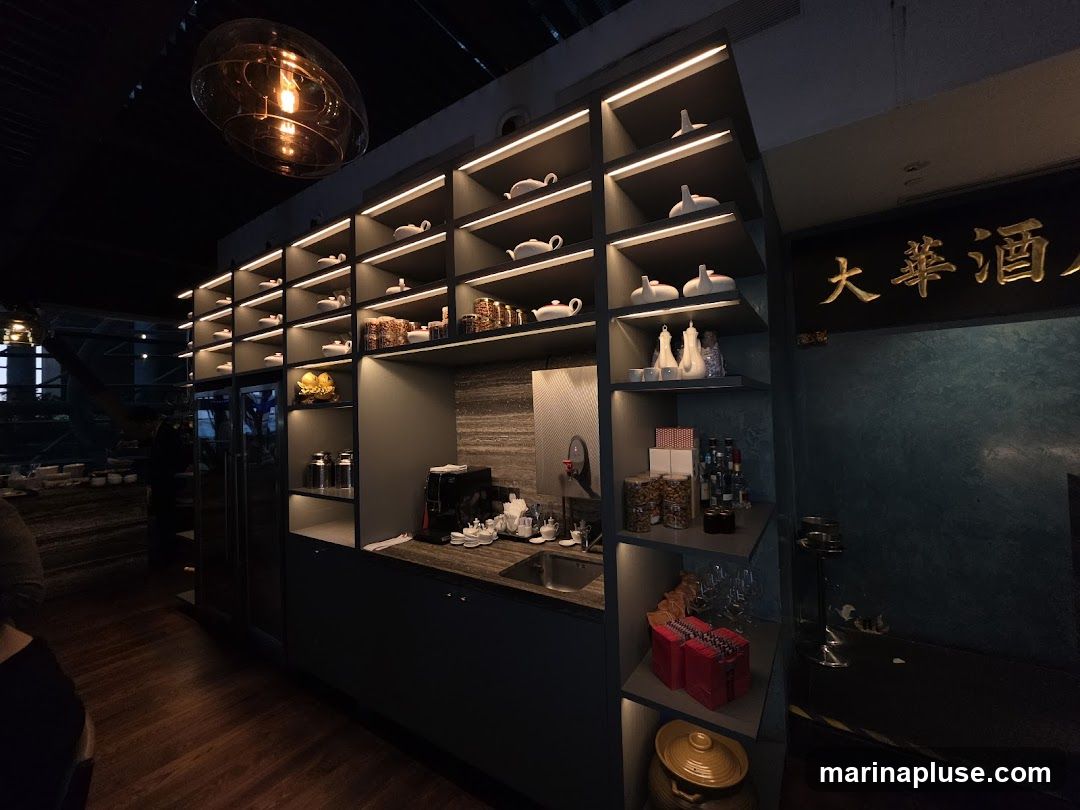 The image shows the interior of a restaurant with a large display unit featuring multiple shelves. The shelves are illuminated and showcase various items, including white ceramic teapots and cups, jars of food, bottles of liquor, and decorative objects. A coffee machine and sink are visible on a counter below the shelves. A large golden sign with Chinese characters is mounted on the wall to the right. The floor is made of dark wood planks.