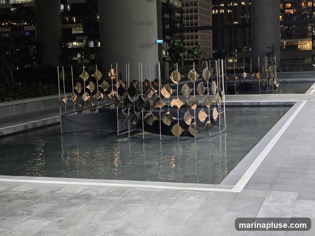 An outdoor water feature at night, with a modern sculptural element made of metallic discs arranged on a frame. The water is calm, reflecting the lights from the surrounding buildings. The area is paved with large grey tiles.