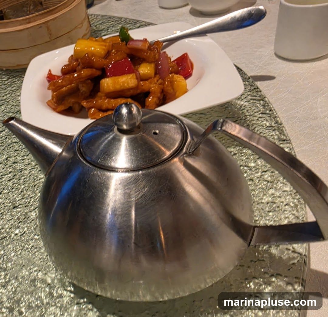A close-up shot of a metal teapot in the foreground, with a plate of sweet and sour pork with pineapple and bell peppers in the background. A bamboo steamer basket is partially visible to the left.