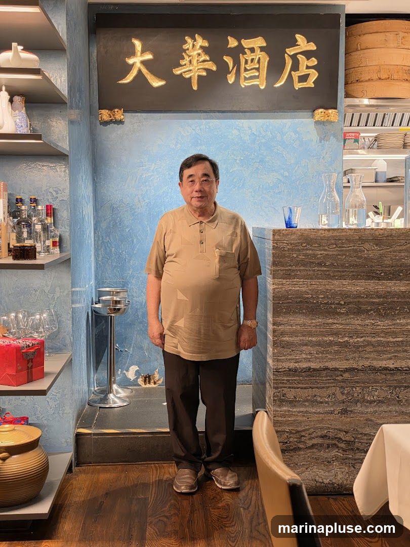 A man stands in what appears to be a restaurant. Above him, a sign with golden Chinese characters is mounted on a blue textured wall. To his left, shelves display bottles of alcohol and decorative items. To his right, a counter area is visible with carafes of water and glassware. A table with a white tablecloth is set in the foreground. The flooring is dark wood.
