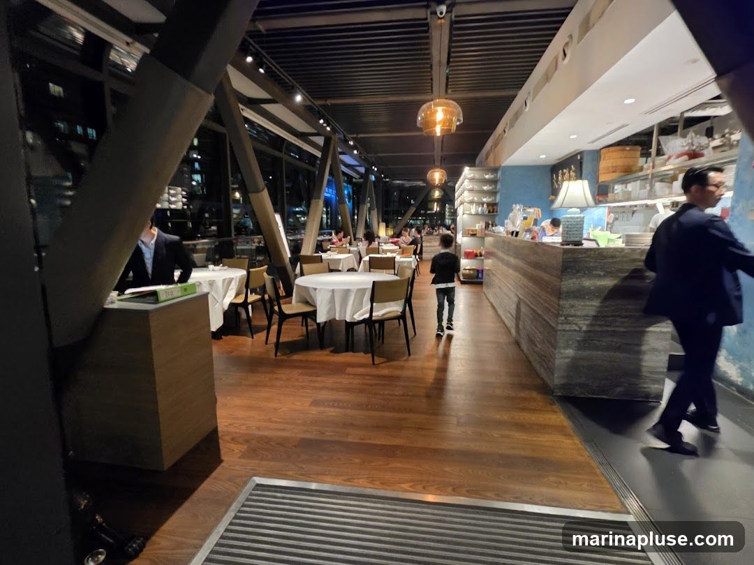 The image shows the interior of a restaurant with tables set for dining. A man in a suit is walking past the counter, and a child is walking towards the counter. The restaurant has a modern design with wooden floors and large windows.