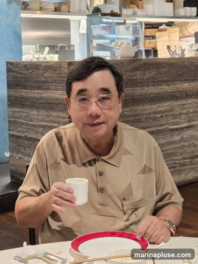 A man with glasses is sitting at a table in a restaurant, holding a white cup in his right hand. He is wearing a tan polo shirt. In the background, there is a display case with food items and a teapot. The table has plates and napkins.