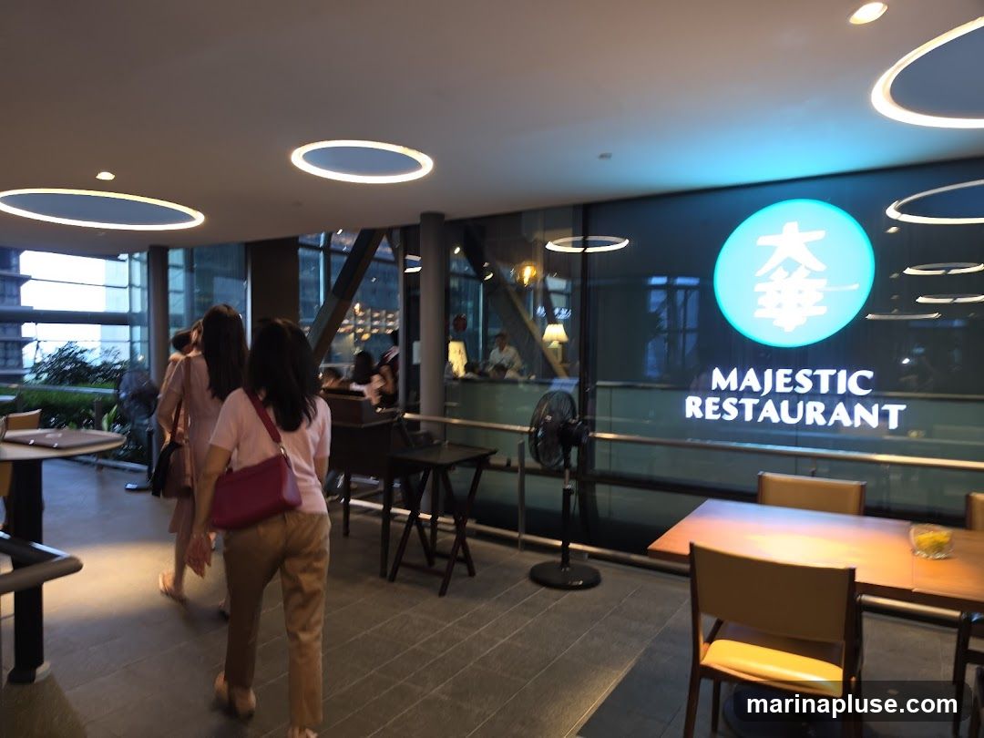 The interior of a restaurant with a large window displaying the "MAJESTIC RESTAURANT" logo. Several tables and chairs are visible, along with people walking inside. The lighting is modern with circular ceiling fixtures.