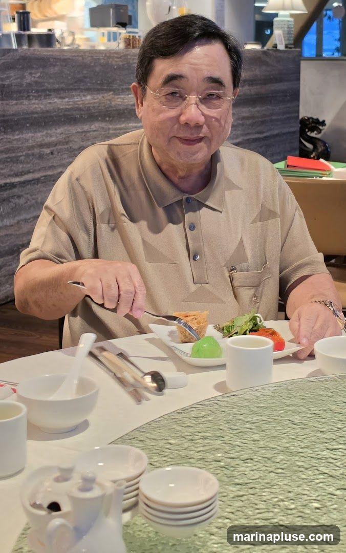 A man with glasses and a tan polo shirt is seated at a table, holding a fork and knife. He is about to eat from a plate containing a small pastry, a green jelly, and a piece of red food with salad. The table is set with white bowls, plates, and teaware. The background shows a restaurant interior with decorative stone walls and a lamp.