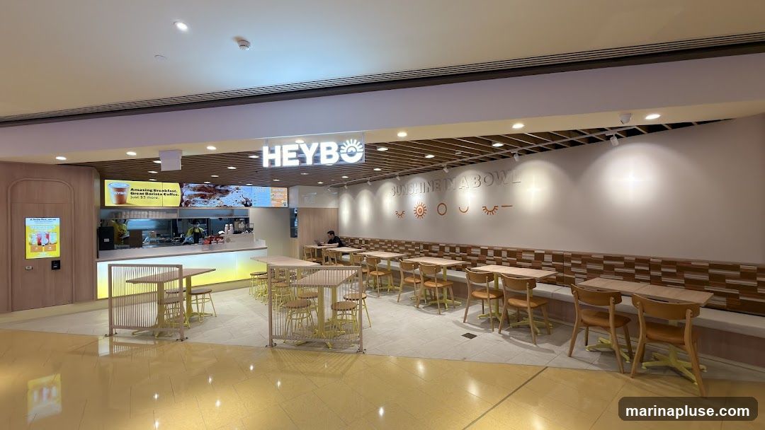 The exterior of a HEYBO shop with a bright, modern interior. The shop features a long counter with staff preparing food, and a seating area with tables and chairs. The walls are decorated with the slogan "SUNSHINE IN A BOWL" and playful sun and eye motifs. The overall ambiance is clean and inviting.