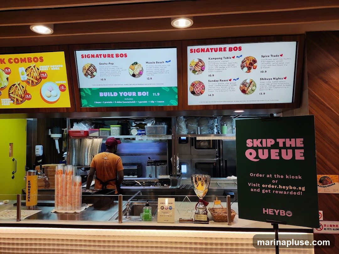 A restaurant counter with digital menus displaying food options and a sign encouraging customers to skip the queue by ordering online. The menu features "Combos," "Signature Bos" with dishes like "Gochu-Pop," "Muscle Beach," "Kampong Table," "Sunday Roast," "Spice Trade," and "Shibuya Nights," as well as a "Build Your Bo!" option. A staff member is visible behind the counter.