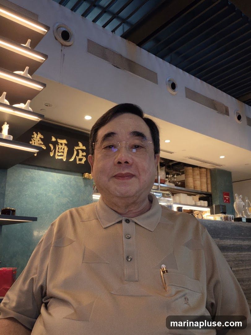 A man wearing a tan polo shirt poses for a photo in what appears to be a restaurant. Behind him, a sign with Chinese characters is visible, along with decorative shelving and what looks like a bar area with steaming baskets in the background.