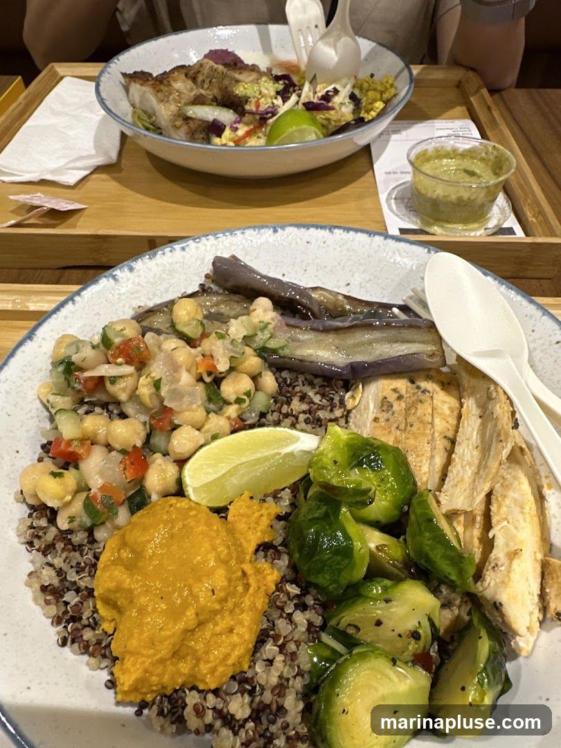 Two bowls of food are presented on wooden trays. The bowl in the foreground contains quinoa, sliced grilled chicken, roasted Brussels sprouts, grilled eggplant, a chickpea salad, and a dollop of orange hummus. The bowl in the background appears to have chicken, salad, and grains. A small container of green sauce is also visible.