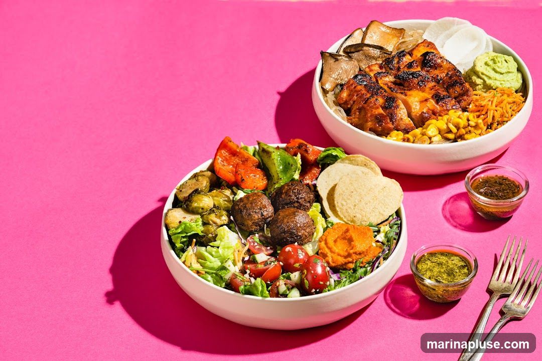 Two white bowls filled with various ingredients are displayed against a vibrant pink background. The bowl in the foreground contains a salad base topped with lettuce, cherry tomatoes, cucumber, roasted Brussels sprouts, red and green bell peppers, meatballs, tortilla chips, and a dollop of orange puree. The bowl in the background features grilled chicken, mushrooms, shredded carrots, corn, and a green puree. Two small glass dishes of dipping sauce and two forks are also visible.