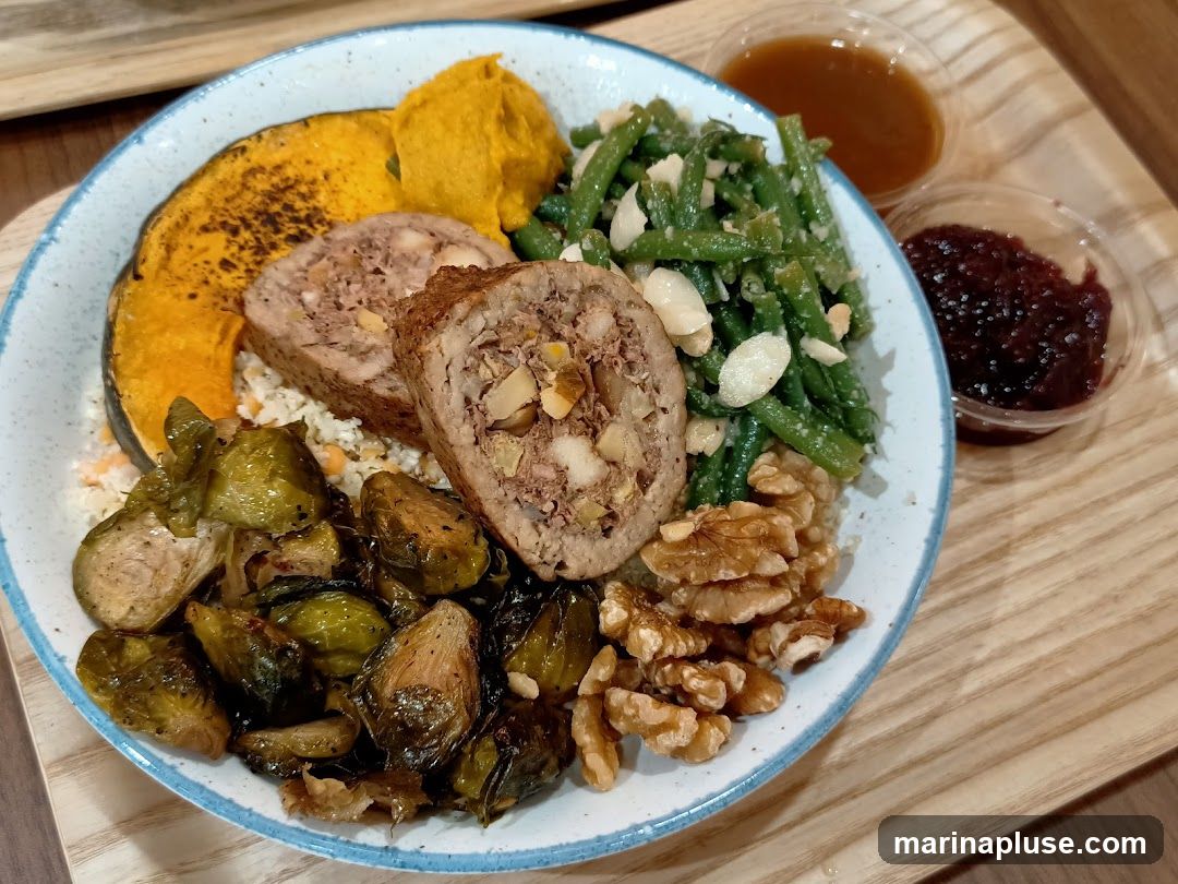 A close-up shot of a meal served in a bowl. The meal includes sliced meatloaf with nuts, roasted Brussels sprouts, a slice of roasted pumpkin, mashed pumpkin, and green beans with almonds. Two small containers of sauce, one dark brown and one red, are on the side. The food is presented on a light wooden tray.
