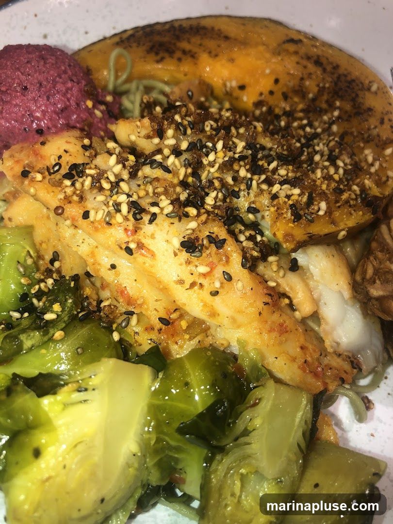 A close-up shot of a bowl of food. The dish contains a piece of white fish seasoned with sesame seeds and spices, roasted Brussels sprouts, a scoop of purple dip, and a slice of roasted squash or sweet potato. Green noodles are visible underneath the other ingredients.