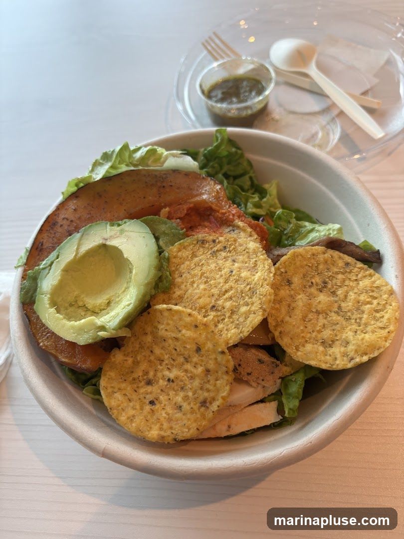 A close-up overhead view of a salad bowl filled with various ingredients. The salad includes lettuce, sliced avocado, roasted sweet potato, shredded chicken, and tortilla chips. A small container of green dressing and disposable cutlery are visible in the background.