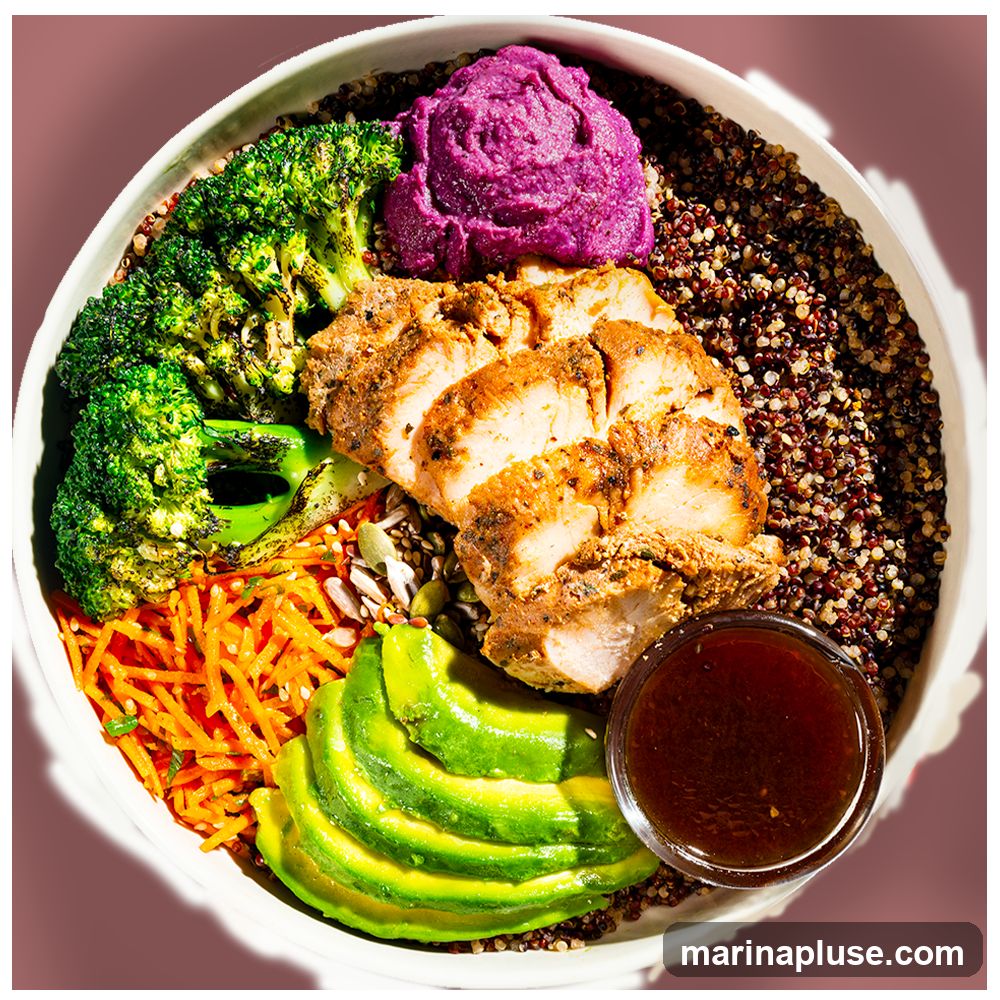 A top-down view of a healthy quinoa bowl. The bowl contains sliced grilled chicken breast seasoned with spices, roasted broccoli florets, shredded carrots, sliced avocado, a dollop of purple mashed sweet potato, and a side of dark dressing in a small glass bowl. The base of the bowl is a mix of red and brown quinoa, sprinkled with seeds.