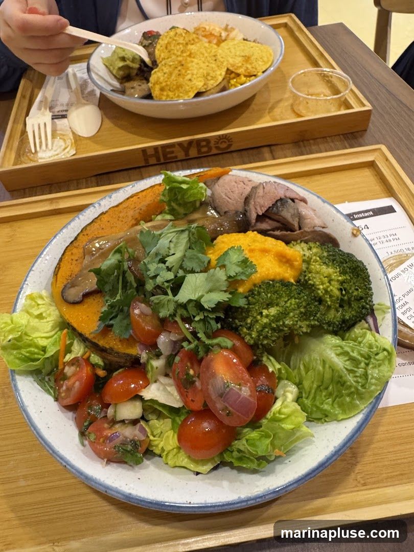 Two bowls of food are shown on wooden trays. The bowl in the foreground contains a salad with lettuce, cherry tomatoes, cucumber, broccoli, roasted pumpkin, sliced meat, and a dollop of orange puree. The bowl in the background appears to have nachos, guacamole, and other ingredients. The word "HEYBO" is visible on the tray in the background. A receipt or menu is partially visible on the right side of the foreground tray.
