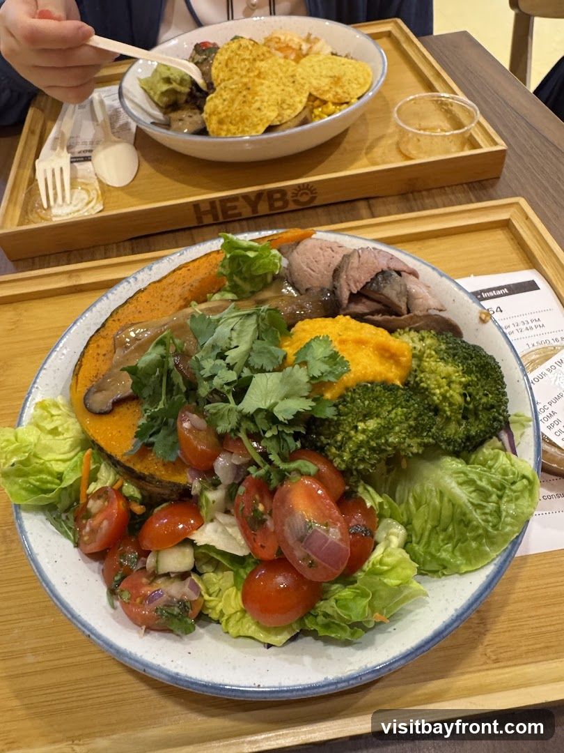 Two bowls of food are shown on wooden trays. The bowl in the foreground contains a salad with lettuce, cherry tomatoes, cucumber, broccoli, roasted pumpkin, sliced meat, and a dollop of orange puree. The bowl in the background appears to have nachos, guacamole, and other ingredients. The word "HEYBO" is visible on the tray in the background. A receipt or menu is partially visible on the right side of the foreground tray.