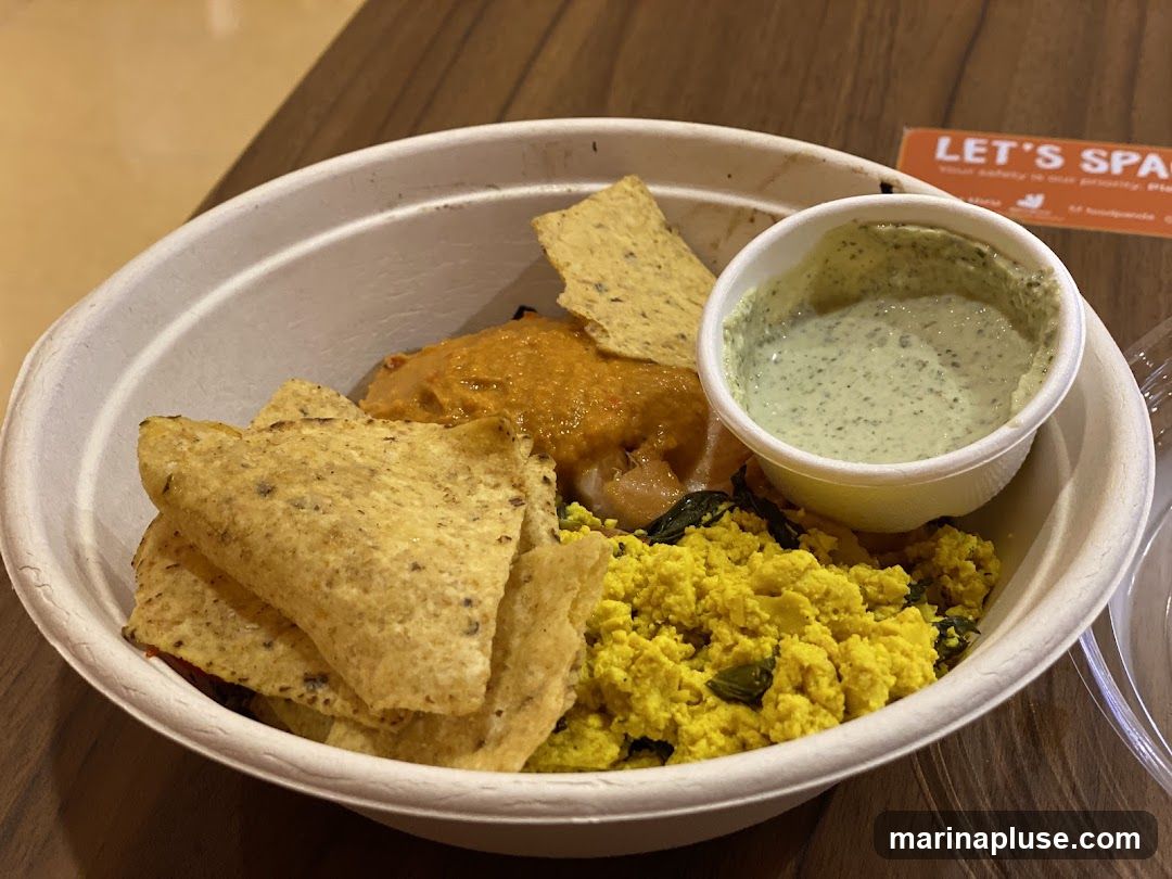 A white bowl filled with a meal. The bowl contains a yellow scramble, possibly tofu, mixed with greens. There are also tortilla chips, a dollop of orange sauce, and a small cup of green sauce. In the background, an orange sign with white text reads "LET'S SPACE".