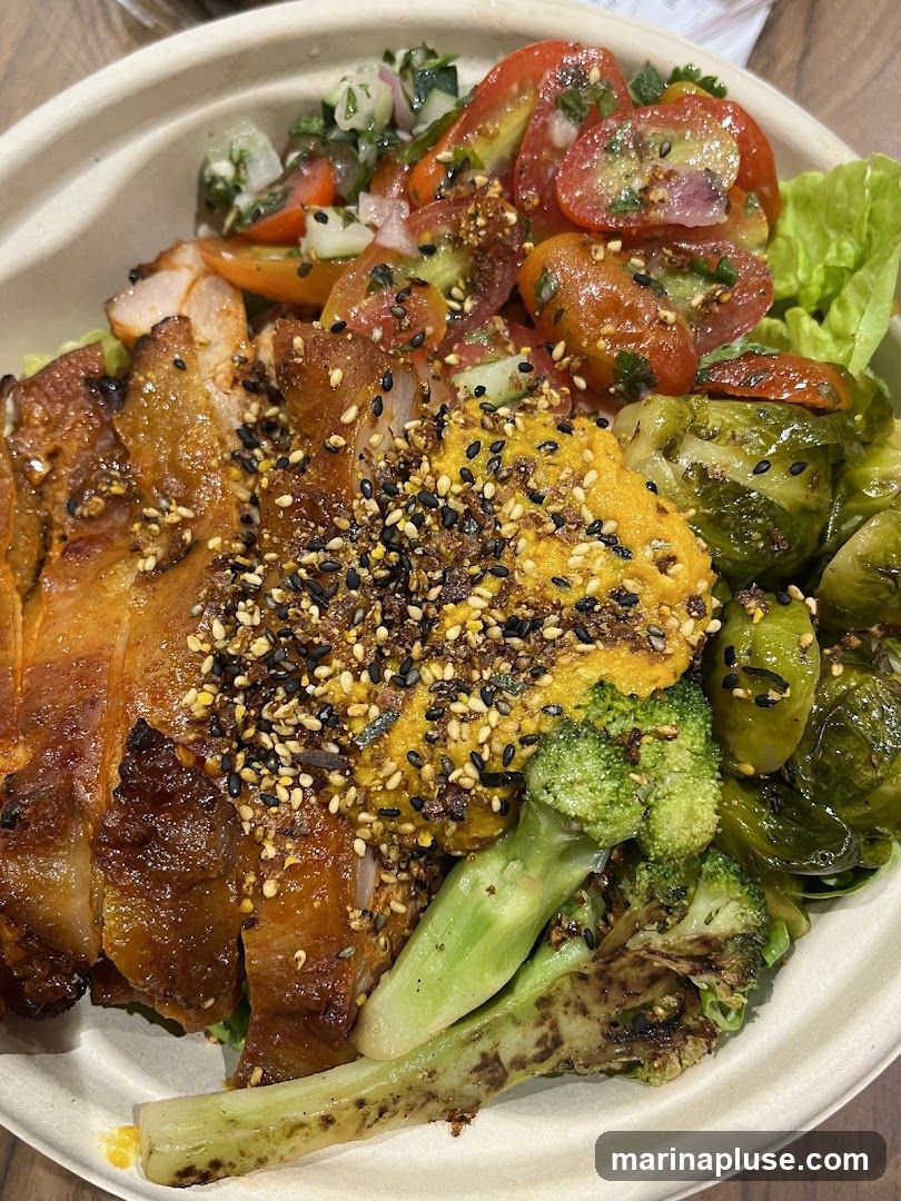 A close-up view of a bowl filled with food. The bowl contains sliced grilled chicken, cherry tomatoes, cucumber, red onion, lettuce, Brussels sprouts, and broccoli. The chicken and vegetables are seasoned with sesame seeds and other spices. The food is served in a disposable bowl.