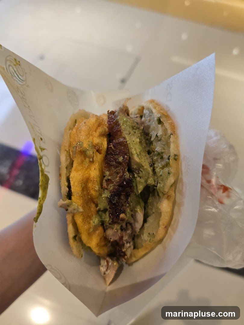 A close-up shot of a half-eaten arepa, a cornmeal cake split open and filled with various ingredients. The fillings appear to include shredded meat, a creamy green sauce, and a yellowish sauce. The arepa is wrapped in white paper with a subtle logo.