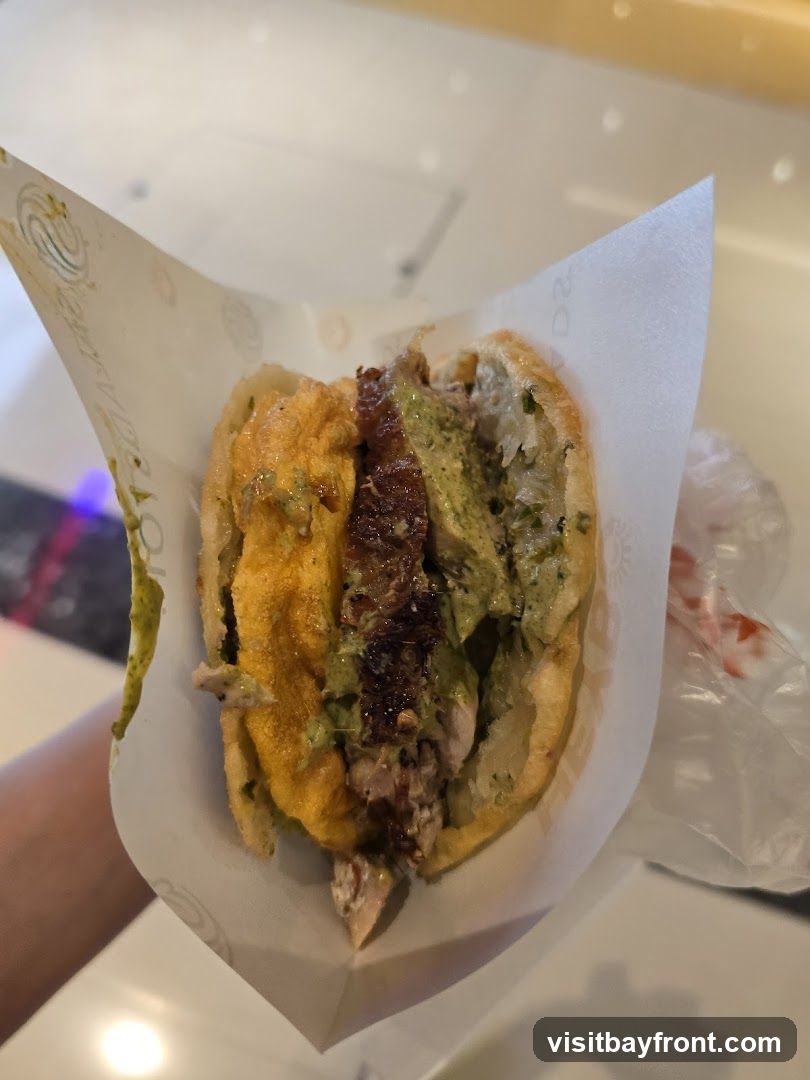 A close-up shot of a half-eaten arepa, a cornmeal cake split open and filled with various ingredients. The fillings appear to include shredded meat, a creamy green sauce, and a yellowish sauce. The arepa is wrapped in white paper with a subtle logo.