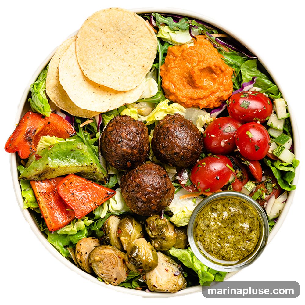 A top-down view of a salad bowl filled with various ingredients. The bowl contains lettuce, red cabbage, roasted Brussels sprouts, grilled bell peppers, cherry tomatoes, cucumber, and three large meatballs. There are also three tortilla chips and a dollop of orange dip. A small glass bowl of green dressing is on the side.