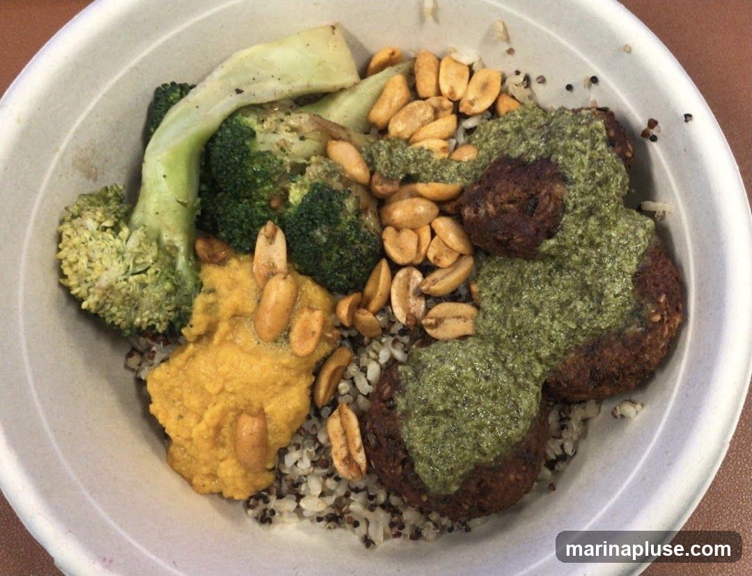 A close-up overhead view of a falafel bowl. The bowl contains a base of quinoa and rice, topped with falafel patties drizzled with a green sauce, a dollop of yellow hummus, roasted broccoli florets, and chopped peanuts.