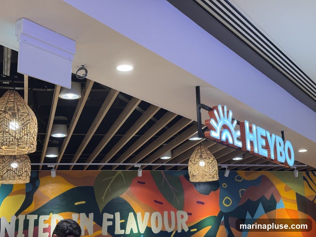 The image shows the interior of a restaurant with a brightly lit sign that says "HEYBO" with a sun graphic. Woven pendant lights hang from the ceiling, and a colorful mural with abstract shapes and leaves is visible on the wall. Part of a person's head is visible in the lower left corner.