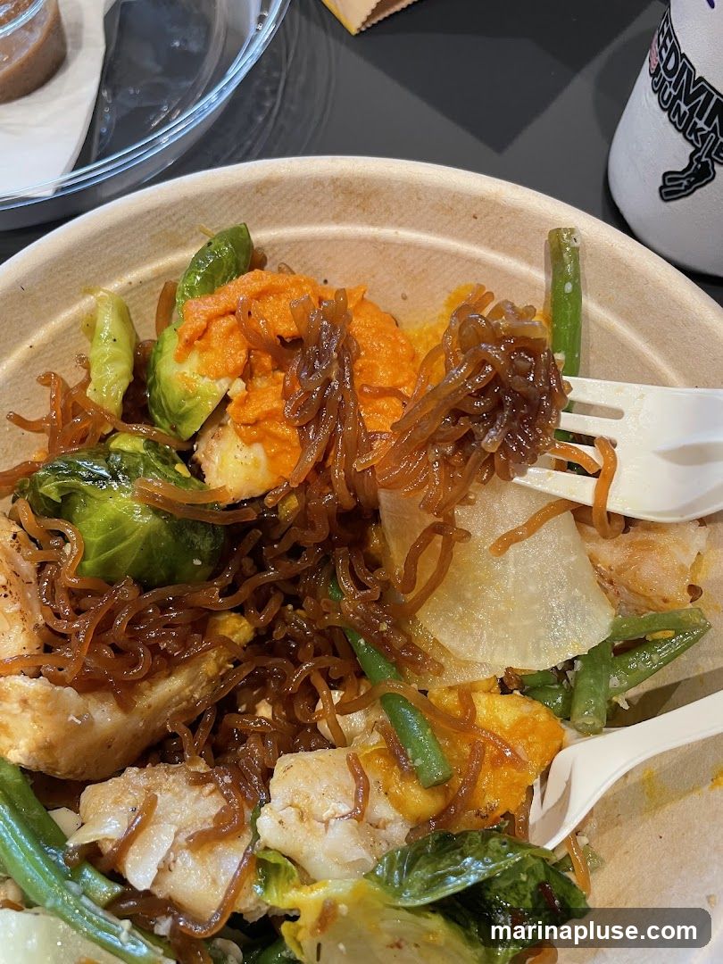 A close-up shot of a bowl filled with food. The dish appears to contain pieces of white fish, Brussels sprouts, green beans, and a sweet potato puree, all mixed with thin, dark noodles. A white fork is visible, partially submerged in the food. In the background, a white cup with black lettering and a clear container with a brown substance can be seen.