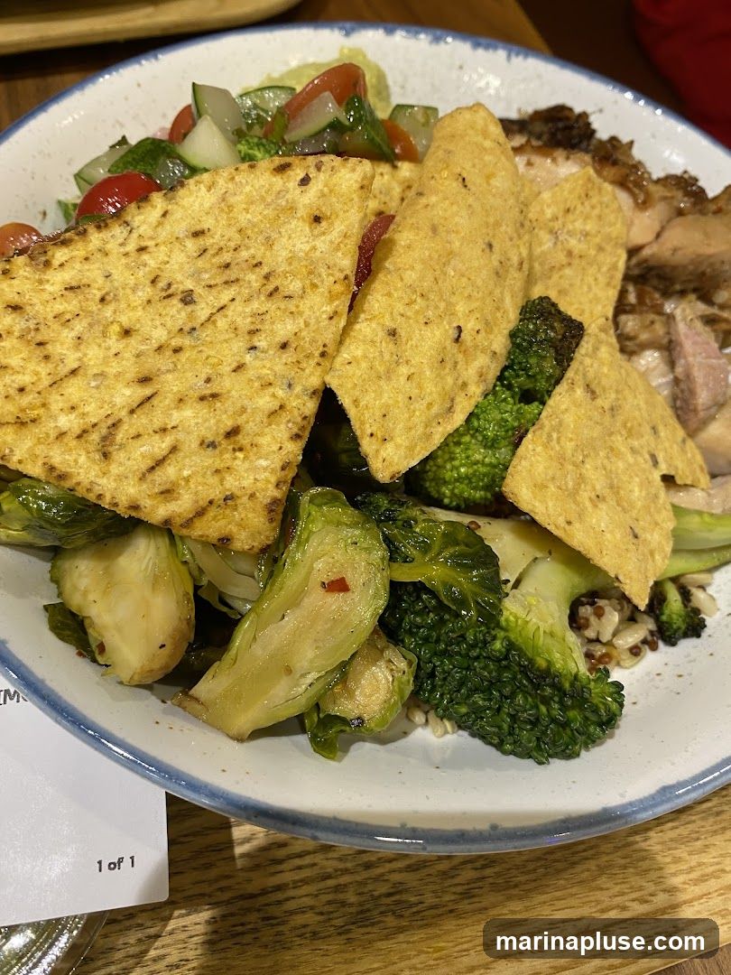 A close-up shot of a salad in a white bowl with a blue rim. The salad contains roasted Brussels sprouts, broccoli florets, cherry tomatoes, cucumber, and tortilla chips. Sliced grilled chicken is visible on the right side of the bowl. A grain base, possibly quinoa or rice, is also present. A small white card with "1 of 1" printed on it is partially visible at the bottom left.