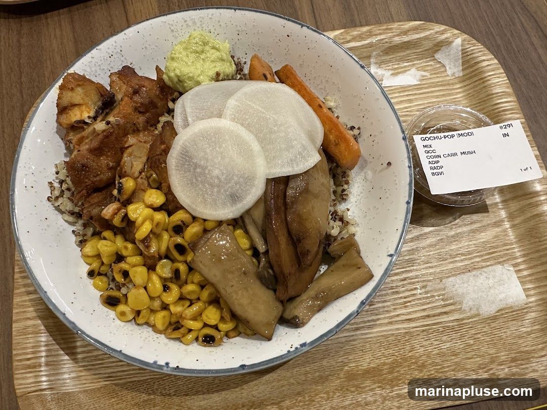 A bowl of food containing chicken, corn, mushrooms, carrots, and radish slices, served with a side of green sauce and a small container of sauce. The bowl is placed on a wooden tray. A label on the side reads "GOCHU-POP [MOD] MIX GCC CORN CARR MUSH ADIP RADP BGVI #291 IN 1 of 1".