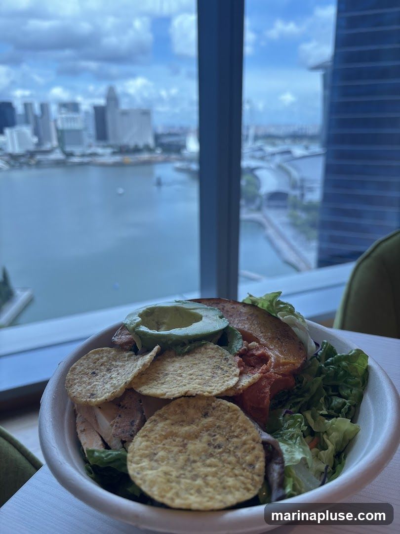 A close-up of a salad in a bowl, featuring chicken, avocado, tortilla chips, and lettuce. The salad is placed on a table in front of a large window with a panoramic view of a city skyline and a body of water. The background is slightly blurred, emphasizing the food in the foreground.