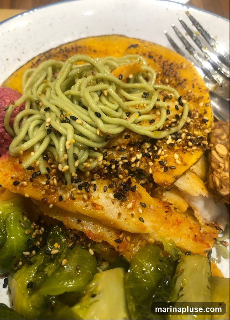 A close-up shot of a healthy bowl dish. The bowl contains pieces of white fish seasoned with spices and sesame seeds, a layer of mashed sweet potato, green noodles, Brussels sprouts, and a dollop of pink hummus. A fork is visible on the right side of the bowl.