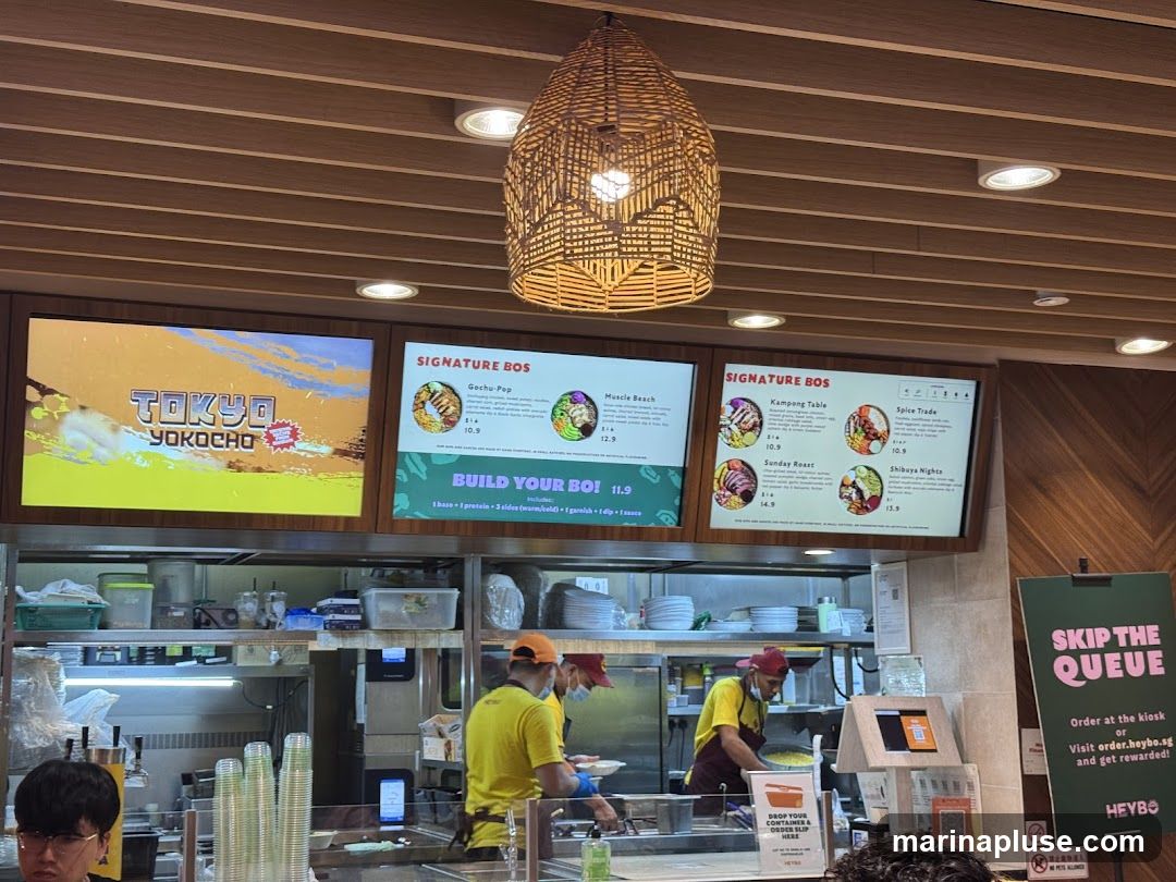 Digital menu boards display food options and prices. One board shows "Tokyo Yokochō" with vibrant graphics. Another board lists "Signature Bos" with items like "Gochu-Pop," "Muscle Beach," "Kampong Table," "Spice Trade," "Sunday Roast," and "Shibuya Nights," along with their prices. Below the menus, staff members are visible working behind a counter. A sign on the right encourages customers to "Skip the Queue" by ordering at a kiosk or online.